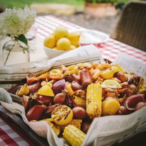 A shrimp boil with shrimp, sausage, corn on the cob, potatoes, onions, and lemon slices served on newspaper, with a bowl of lemons and flowers on a checkered table outdoors.