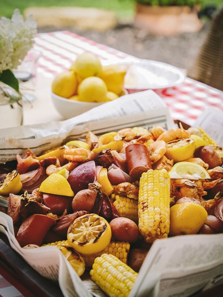 A shrimp boil with shrimp, sausage, corn on the cob, potatoes, onions, and lemon slices served on newspaper, with a bowl of lemons and flowers on a checkered table outdoors.