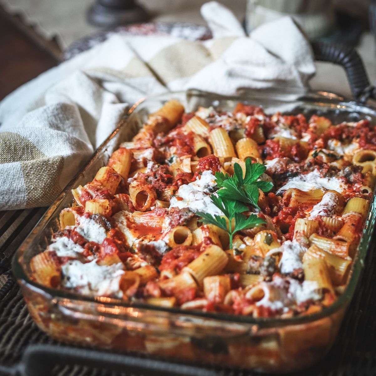 A glass baking dish filled with baked rigatoni pasta in tomato sauce, topped with melted cheese and garnished with fresh parsley. A beige and white kitchen towel is in the background.