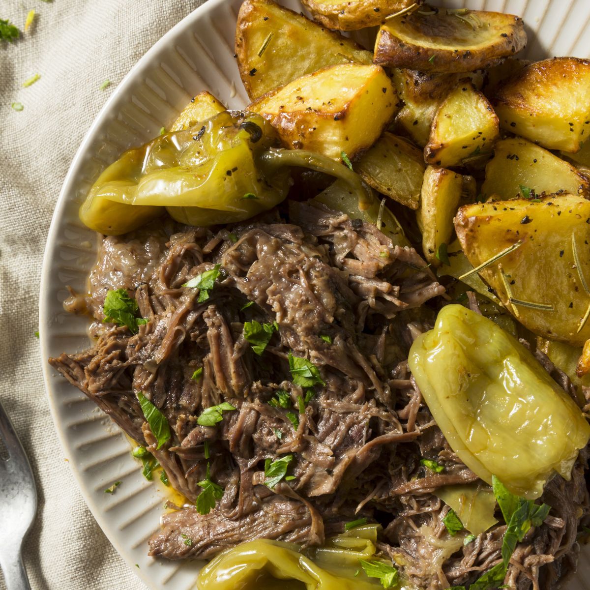 A plate of Mississippi pot roast served with golden roasted potatoes and whole pepperoncini peppers, garnished with chopped parsley. The beef is tender and shredded, surrounded by its savory juices.