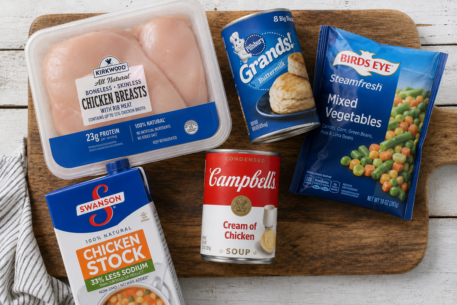 Ingredients for slow cooker, chicken and dumplings with biscuits. It has chicken, biscuits, cream of chicken soup, chicken stock, and mixed vegetables on a wood cutting board.