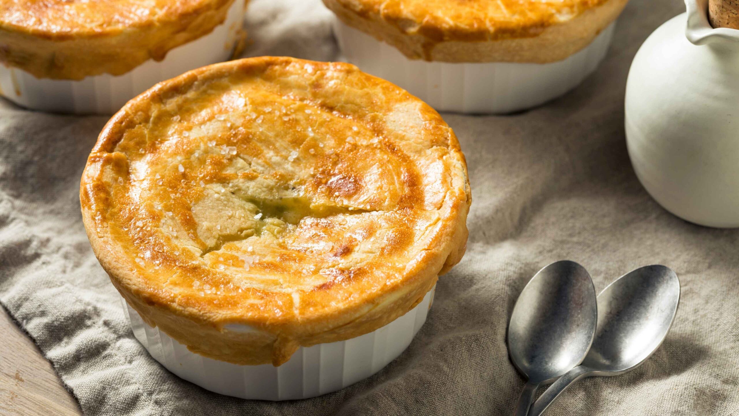 Individual chicken pot pies, and a ramekin on a linen napkin with silver spoons with a golden crust folding over the top with sea salt.