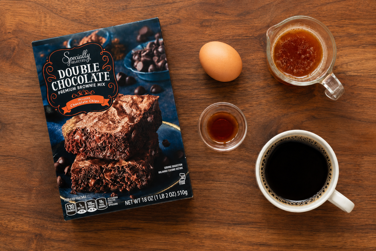 Flat lay of boxed brownie mix, one egg, brown butter, vanilla extract, and black coffee on a walnut cutting board.
