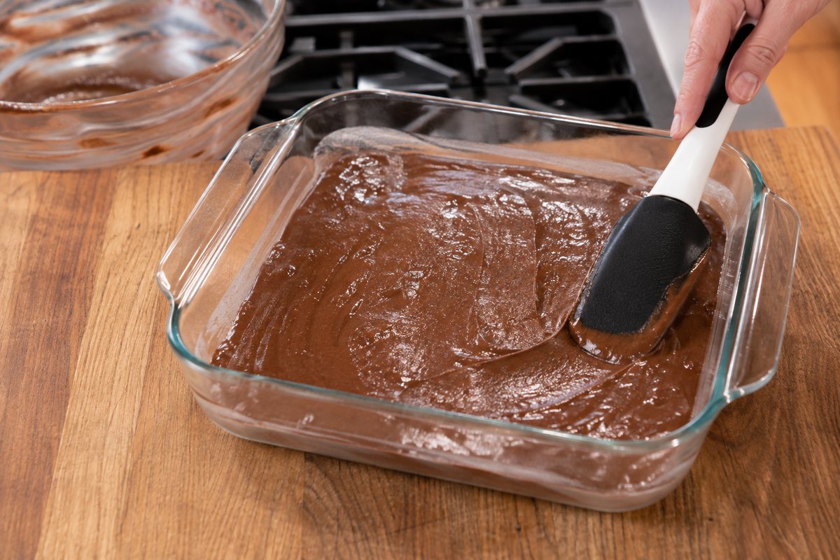 Chocolate brownie batter being spread evenly in an 8x8-inch glass baking pan.