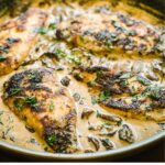 Pan-seared chicken breasts in a creamy mushroom sauce, garnished with fresh herbs. The chicken is browned and surrounded by a rich sauce with pieces of mushrooms and green herbs on top.