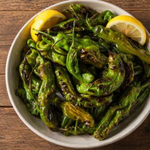 Blistered Shishito peppers in a bowl that are garnished with lemon slices and on a wood table