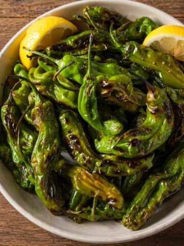 Blistered Shishito peppers in a bowl that are garnished with lemon slices and on a wood table