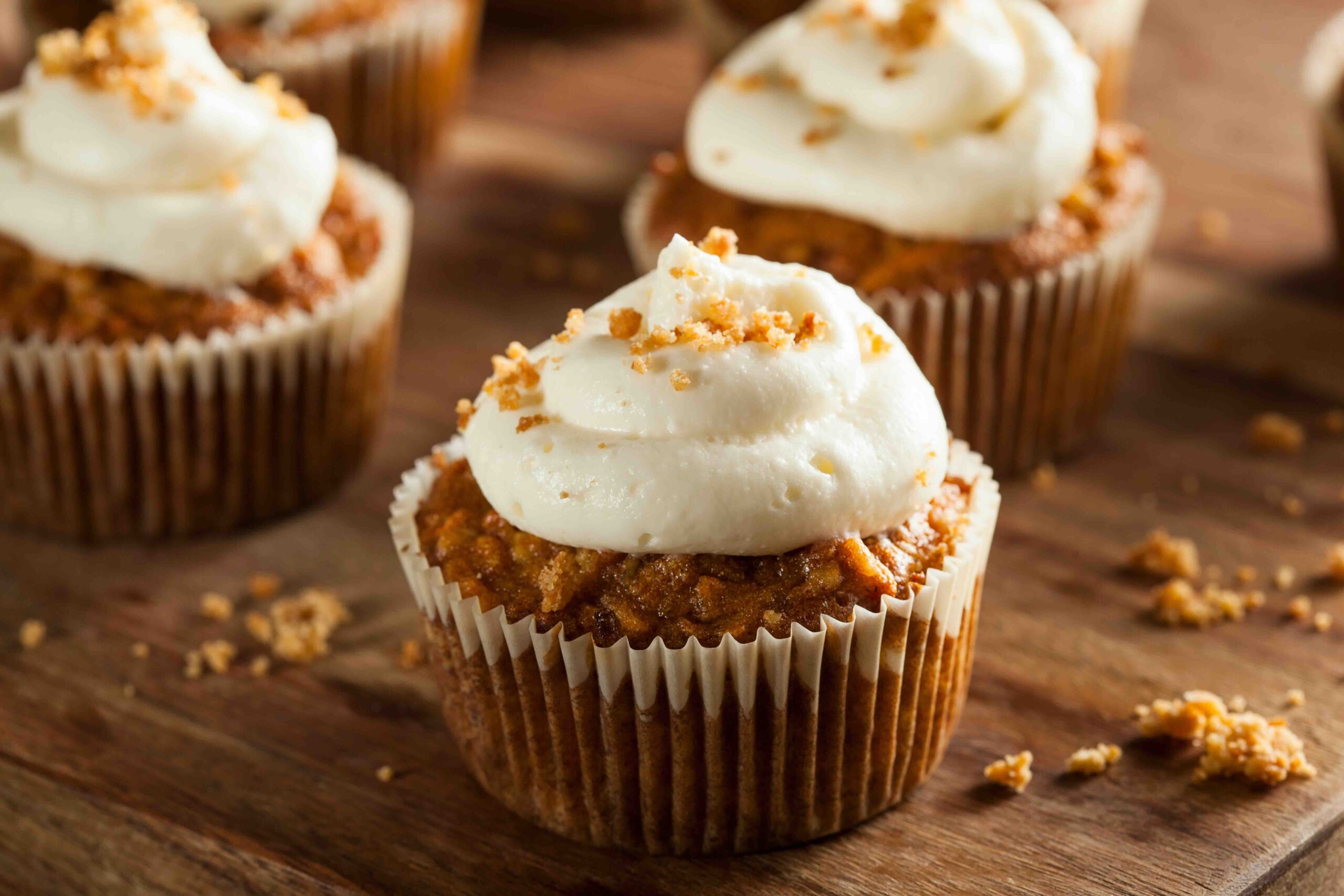 Carrot cake cupcakes with cream cheese frosting on a cutting board with chopped nuts sprinkled on top.