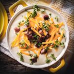 A yellow crock filled with baked potato soup, garnished with bacon Lara's shredded cheddar cheese and parsley. Sitting on a yellow and white tea towel on top of a wooden table.