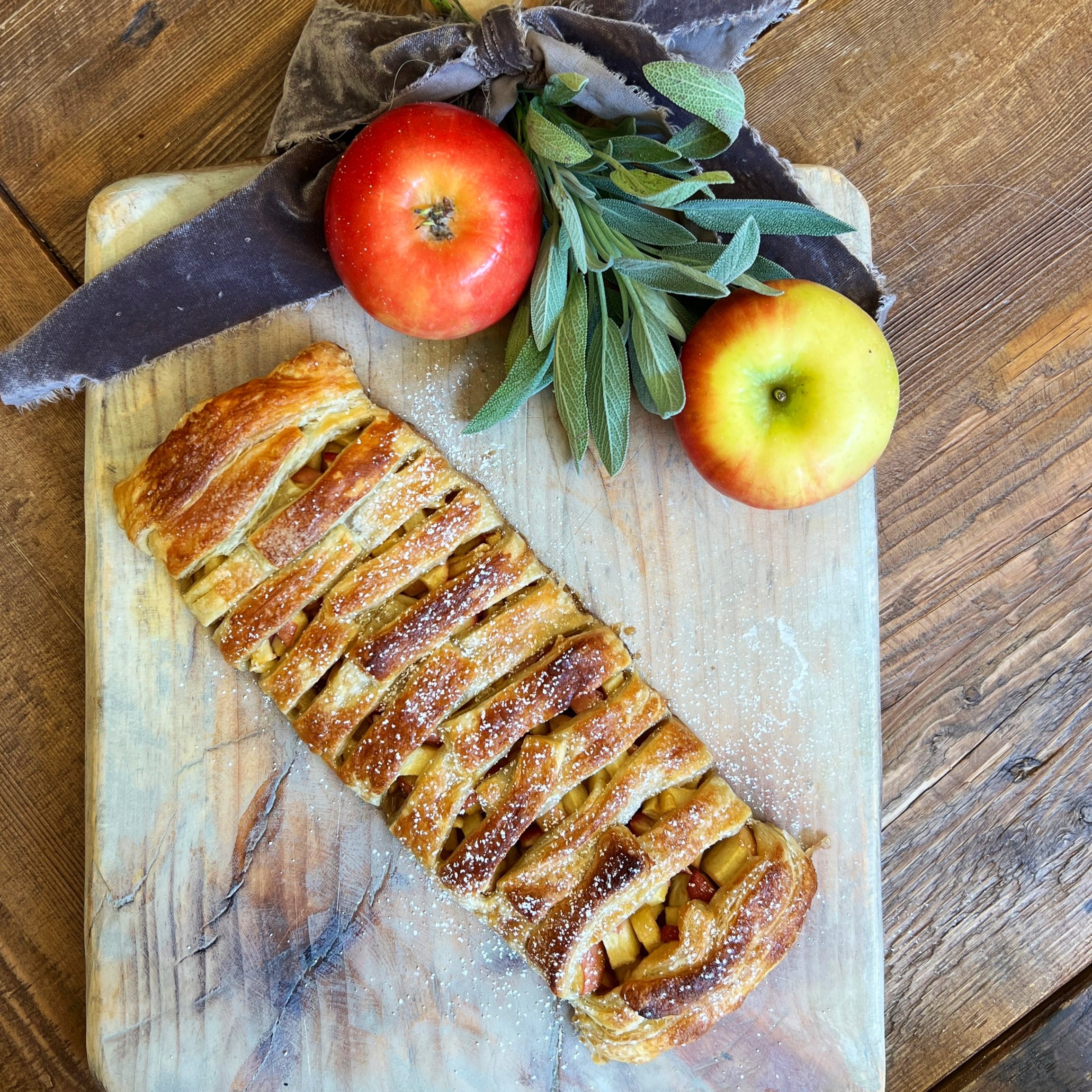 Top of a cutting board with a big velvet ribbon and Sage tucked in as a garnish. Two uncut apples, or else on a cutting board, along with a beautiful golden brown puff pastry filled with apples and topped with the brown sugar garnish.