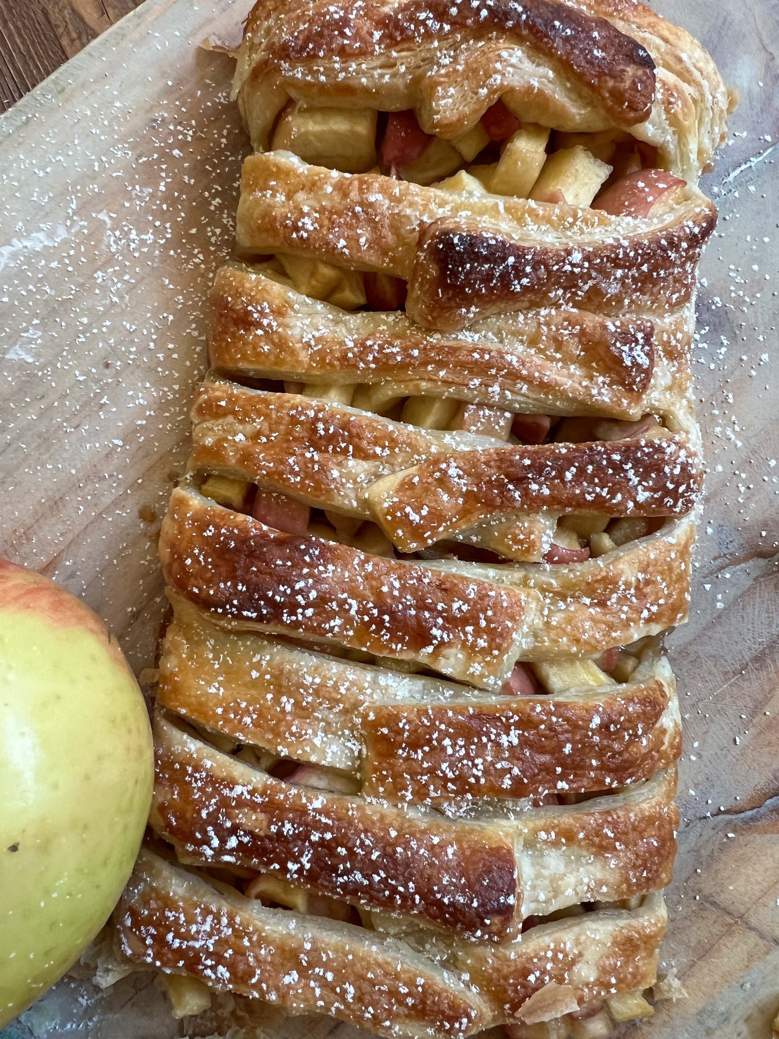 Top view of an apple puff pastry with a lattice top sprinkled with powdered sugar. On a cutting board with an apple as a garnish.