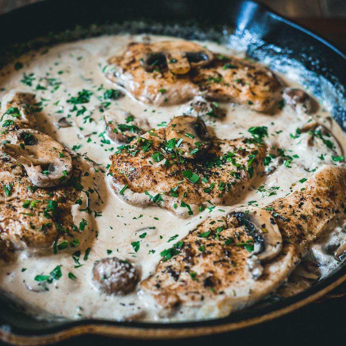 Creamy Parmesan chicken in a skillet, with a Parmesan cream sauce. It has a creamy Parmesan sauce that includes mushrooms and garlic. And the dishes sprinkled with herbs.