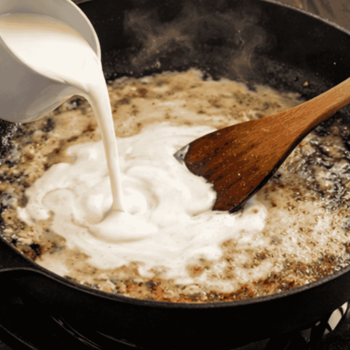 Heavy cream being poured into skillet to create creamy garlic parmesan sauce. Picture of cream and a wooden spoon.